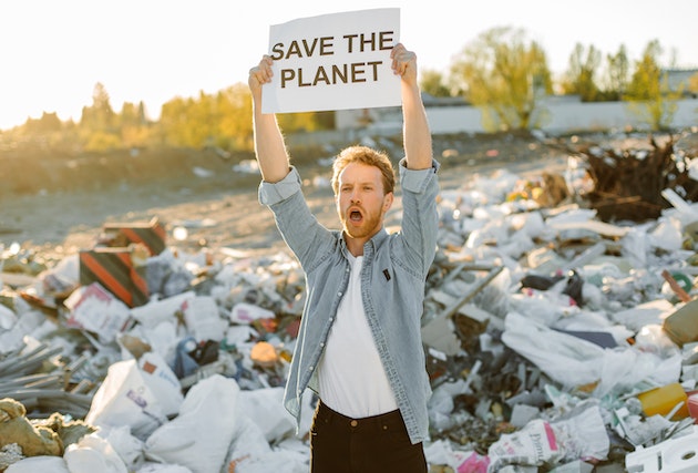 An environmental activist holding a sign with the words "Save the Planet", standing on a beach covered with litter. 