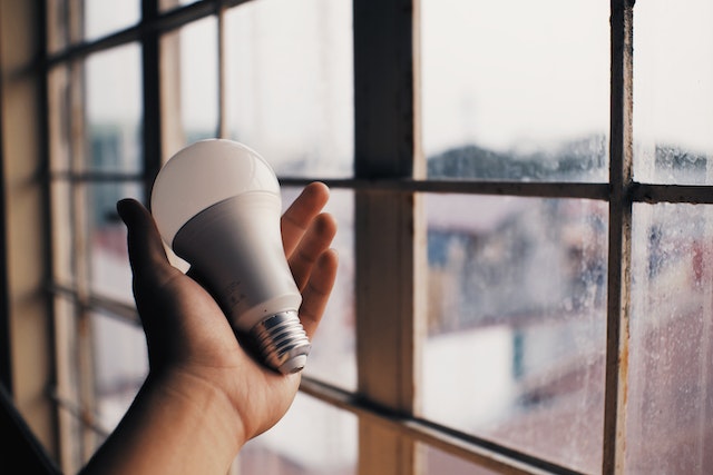 A hand holding an LED bulb against a window. 